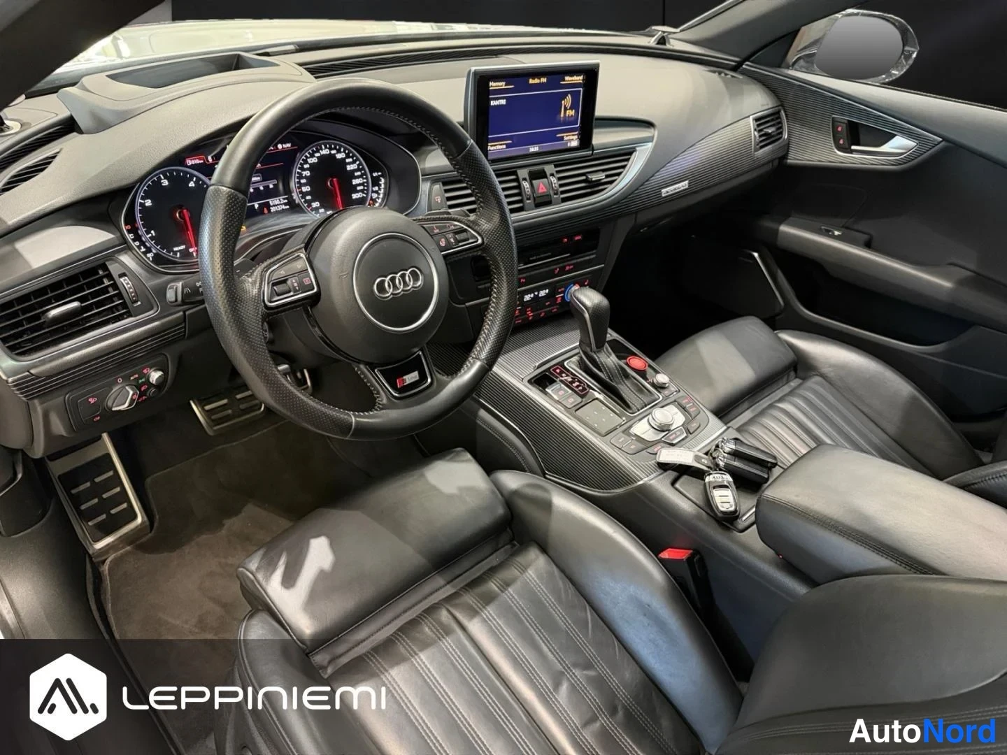 Sivulta: Ohjaamo kulmasta kuvattuna: Audi-ohjauspyörä, mittaristo, keskinäytön infotainment ja mustat nahkapenkit.
