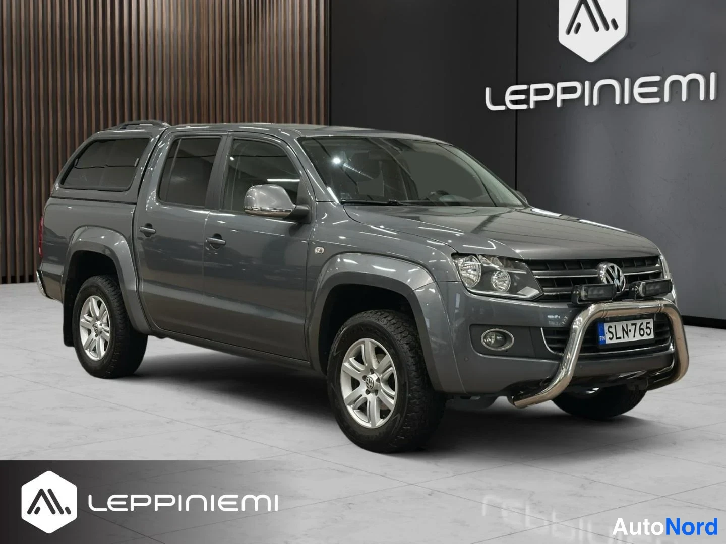Etuviisto, nokka oikealle: Harmaa Volkswagen Amarok -avolava etuokolikulmassa oikealta; etusäleikön Volkswagen-merkki, ajovalot ja A-palkki rekisterillä näkyvät.