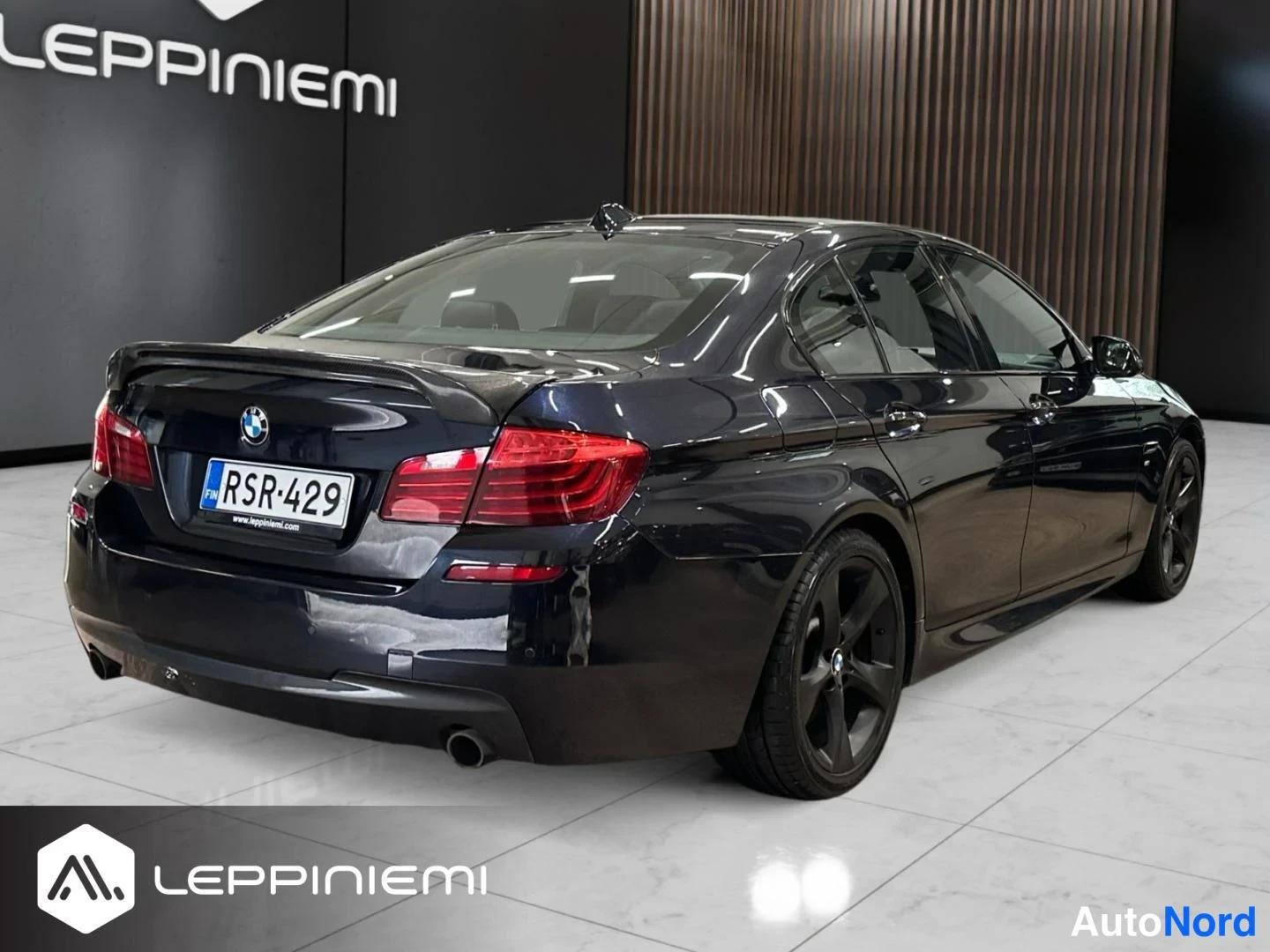 Takaviisto: Takakulmanäkyvä tummansininen BMW 5-sarjan sedan hopeisella Lepanniemen-Leppiniemi-taustalla, rekisterikilpi.