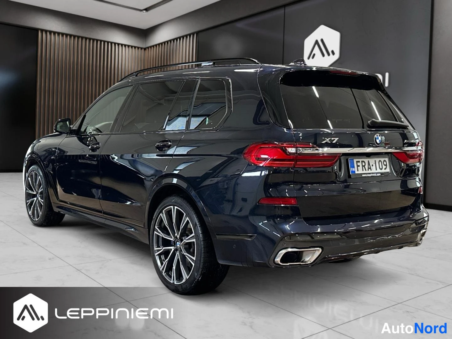 Takaviisto: Taka kolmasosa BMW X7:stä vasemmalta takaa kuvattuna. Tummansininen kori, mukana X7-merkintä, takavalojen kromilista, rekisteritunnus, kattohelinät ja monipaikk