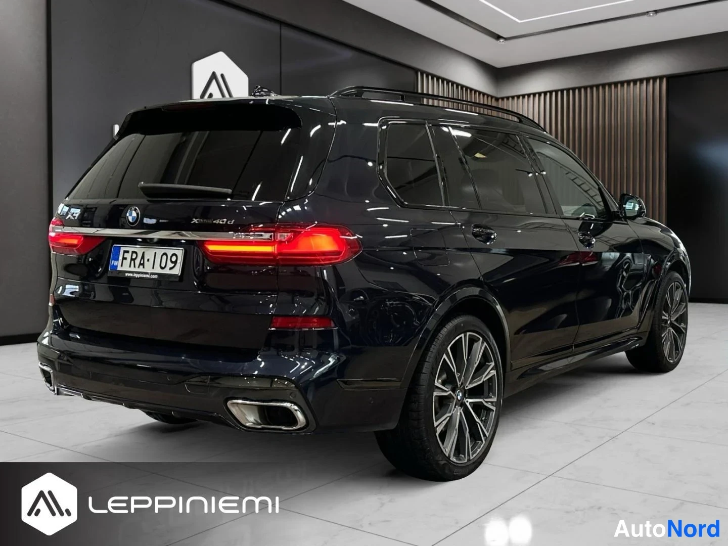 Takaviisto: Tummansininen BMW X7 takaviisto vasemmalta, rekisterikilpi näkyvissä showroomissa.