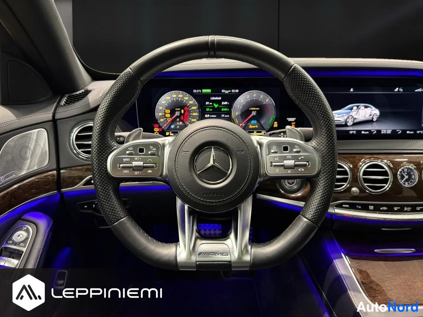 Muu: Lähikuva AMG-ohjauspyörästä Mercedeksessä, taustalla digitaalinen mittaristo ja keskinäyttö, Burmester-kaiuttimen näkyvä osa vasemmalla.