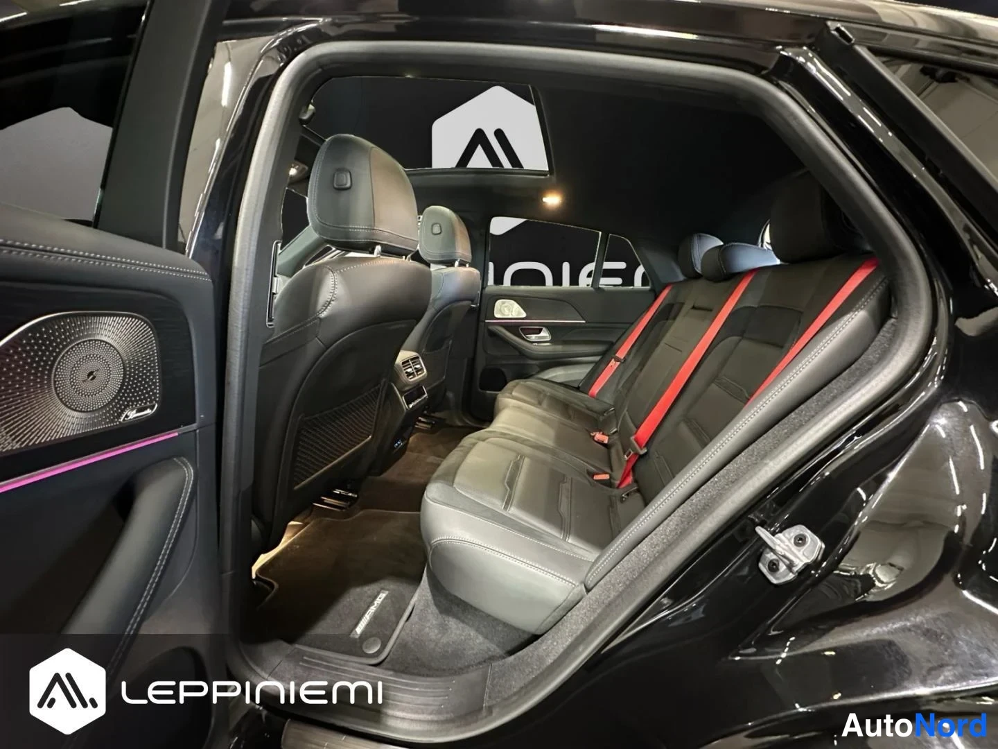 Muu: Mercedes-Benzin takamatkustustila takaoikean oven avattuna: mustat takapenkin istuimet punaisilla turvavyöillä, panoraamakatto, tumma katto ja LEPPIEMI-logon si