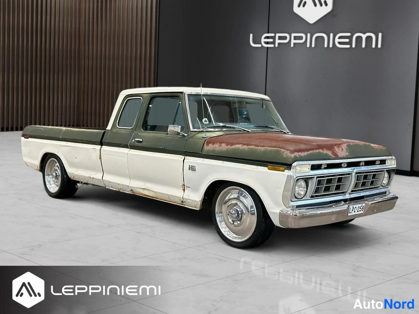 Etuviisto, nokka oikealle: Valkoinen ja tummanvihreä kaksiovinen Ford F-100 lava-auto etukulmasta oikealta, maalipintaa rapautunut konepellissä.