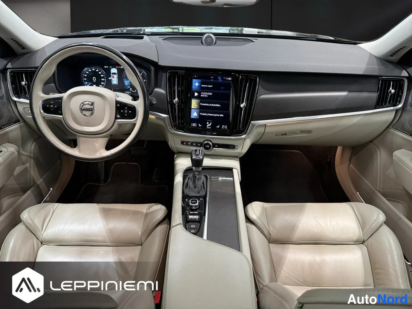 Edestä: Volvo-mallin ohjaamo edestäpäin: kaksi beigeä nahkaistuinta, keskikonsolissa vaihdevalitsin ja infotainment-näyttö.