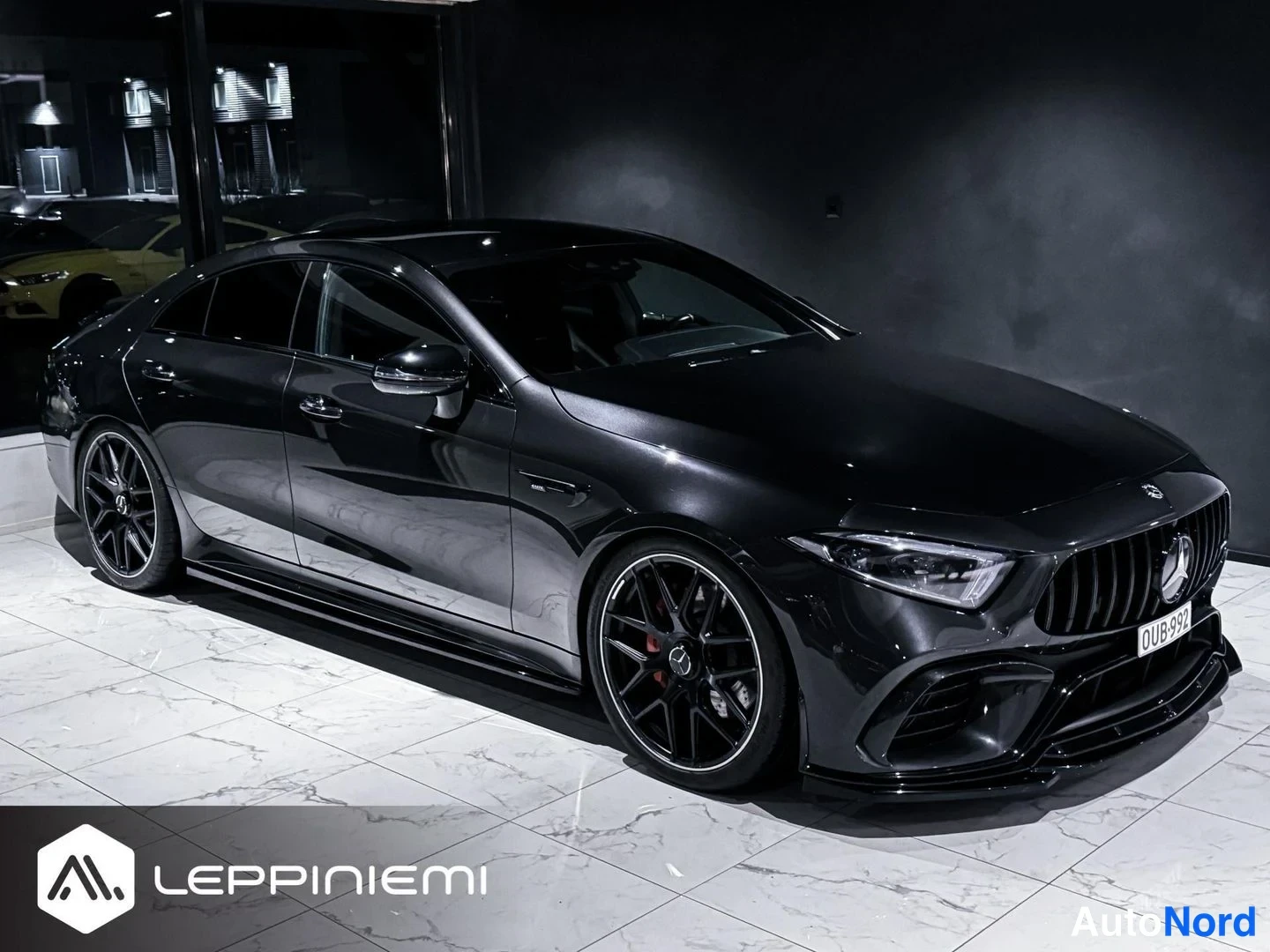 Etuviisto, nokka oikealle: Musta Mercedes-Benz CLS -coupe etukulmasta oikealta sisätiloissa, mustat AMG-vanteet ja punaiset jarrusatulat näkyvät, rekisterikilpi.