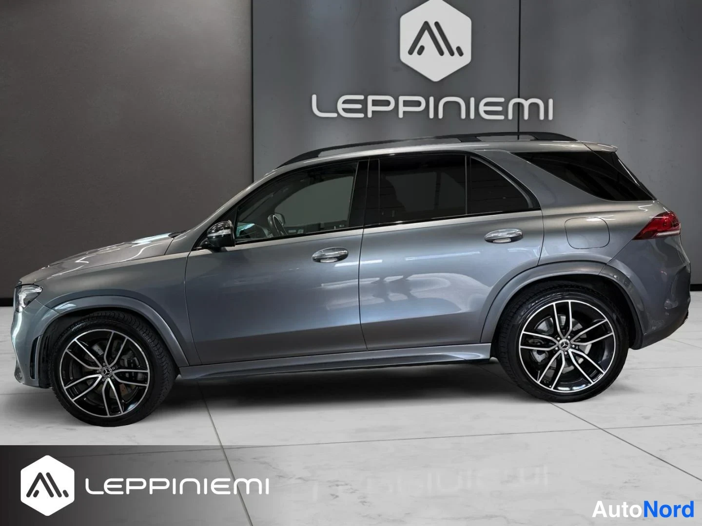 Etuviisto, nokka vasemmalle: Mercedes-Benz GLE:n etukulman vasen näkymä: harmaanmetallivärinen SUV, jonka sivupaneelit, mustat monioksvanteet ja kattokaiteet näkyvät showroomissa.