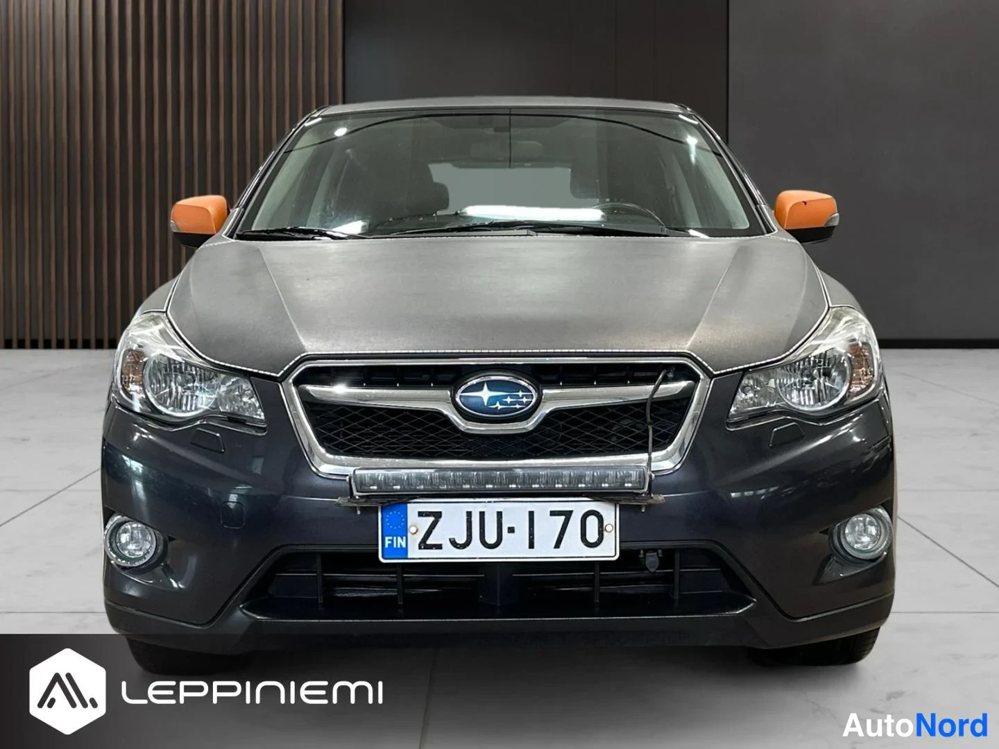 Edestä: Tummanharmaan auton etusivu suoraan edestä, oranssit peilit, Subaru-merkki ja rekisteritunnus.