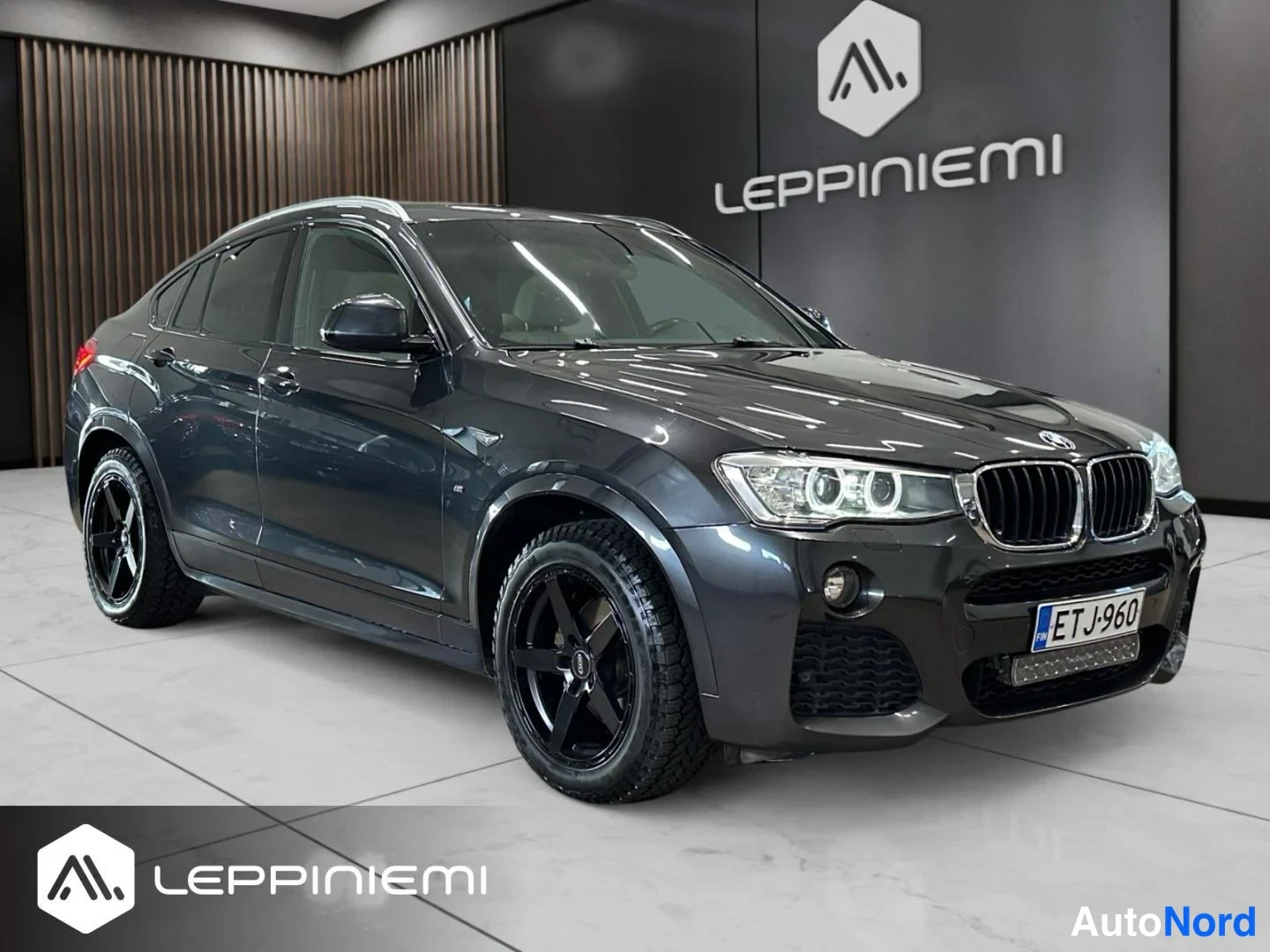 Etuviisto, nokka oikealle: BMW X4:n etukulman kaukokuva sisätiloissa, keula oikealla; tummanharmaa koripinta näkyy, mustat kevytmetallivanteet, sumuvalot ja rekisteritunnus.