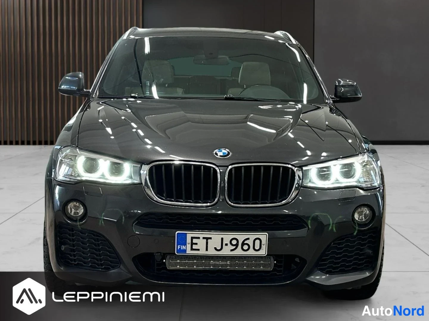 Edestä: Suora etänäkymä BMW X4:stä sisätiloissa; tummanharmaa konepelti, BMW-merkki, etufarot syttyneinä, sumuvalot ja rekisteritunnus.