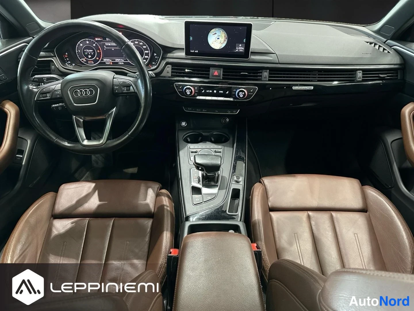 Edestä: Näkymä kojelaudasta ja keskinäytöstä: Audi-ohjauspyörä, kaksi ruskeaa nahkaistuintä, automaattivaihteen valitsin ja kuppipidikkeet keskikonsolissa.