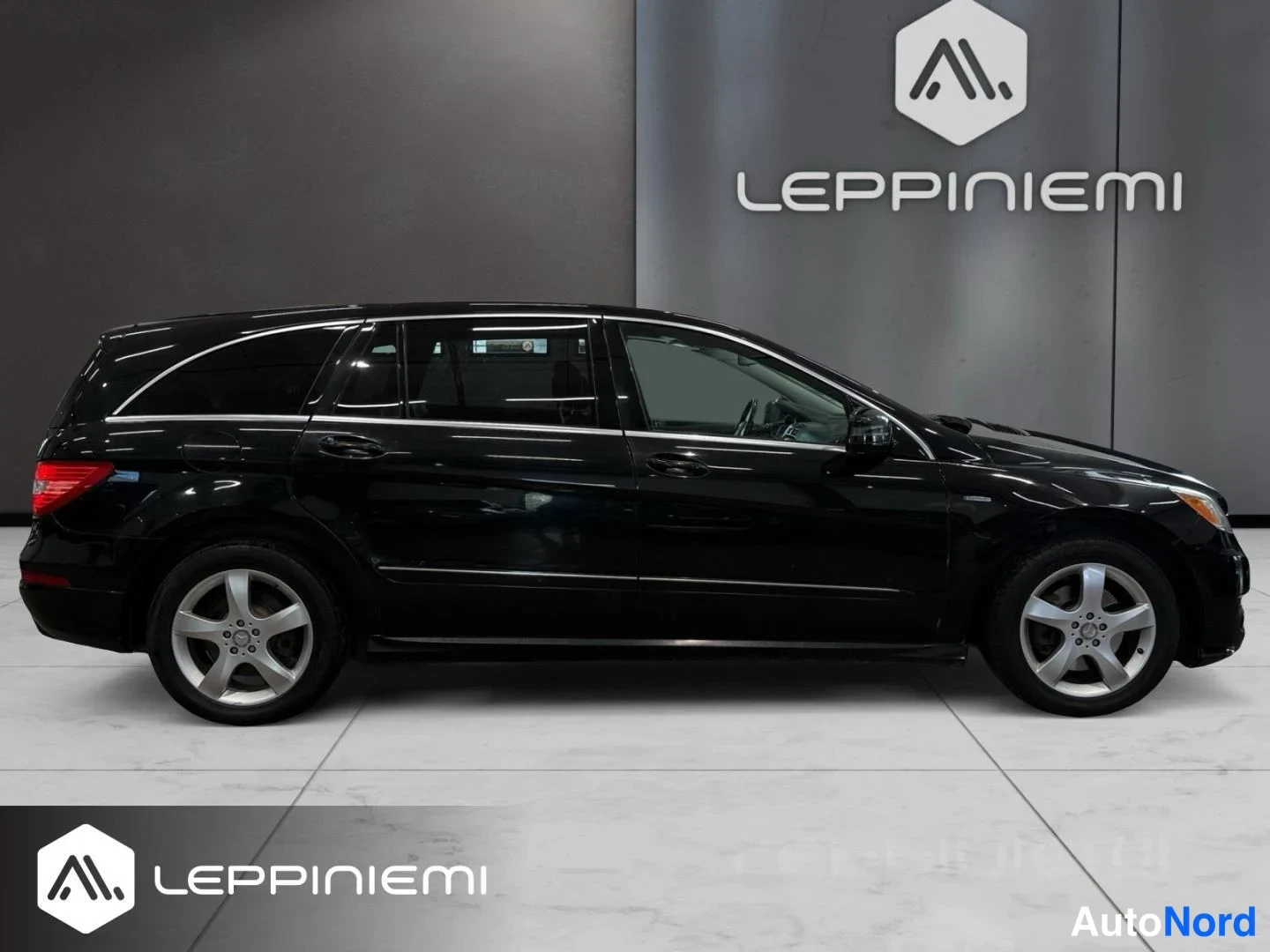 Sivulta: Mustan Mercedes-Benz R -mallin sivunäkymä vasemmalta, tummat ikkunat, kromilista ovissa ja hopeiset viisipuutankkoratteet.