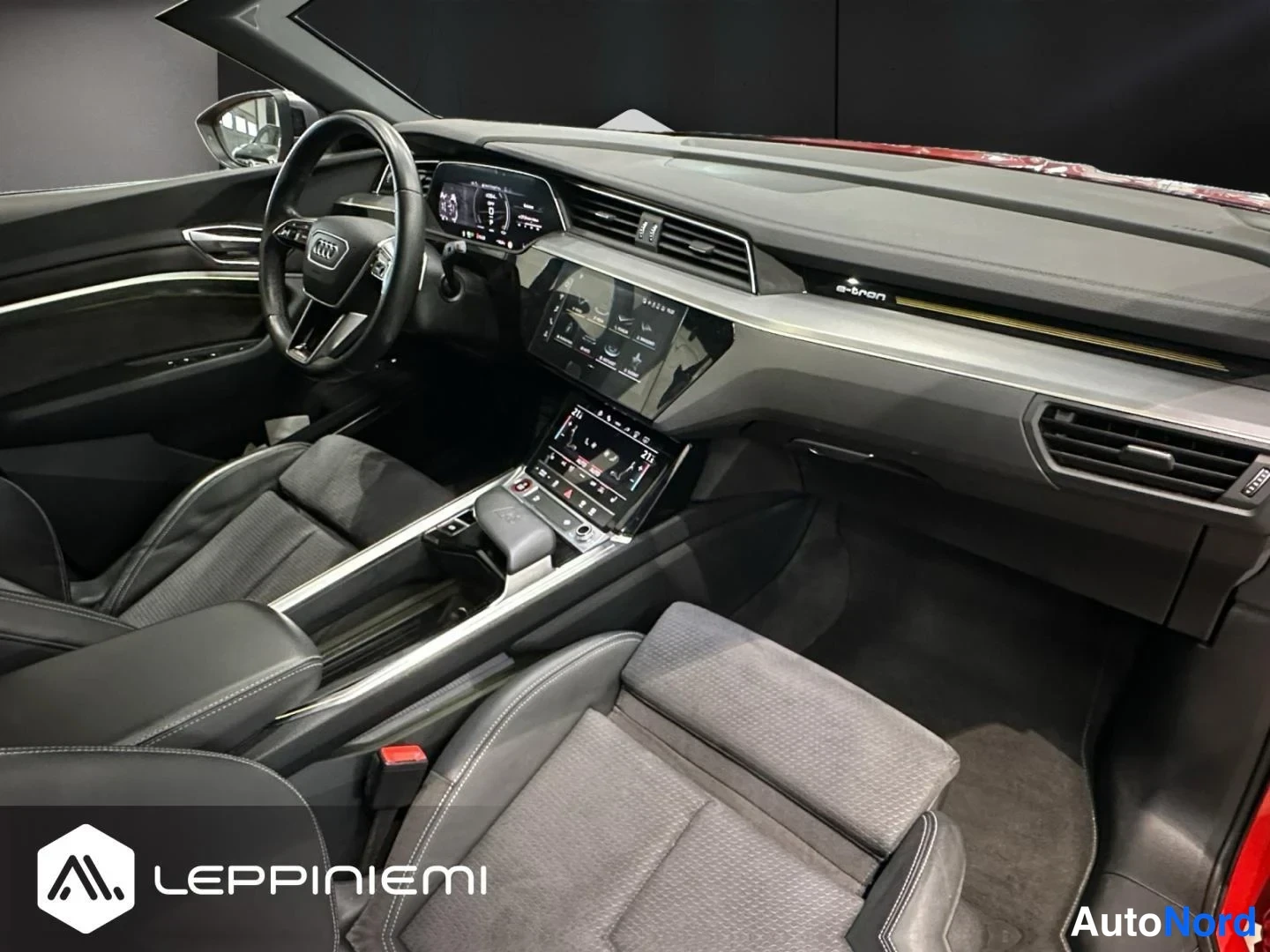 Muu: Audi e-tronin ohjaamo: musta sisusta, Audi-ohjauspyörä, kaksi näyttöä ja e-tron-teksti kojelaudassa.