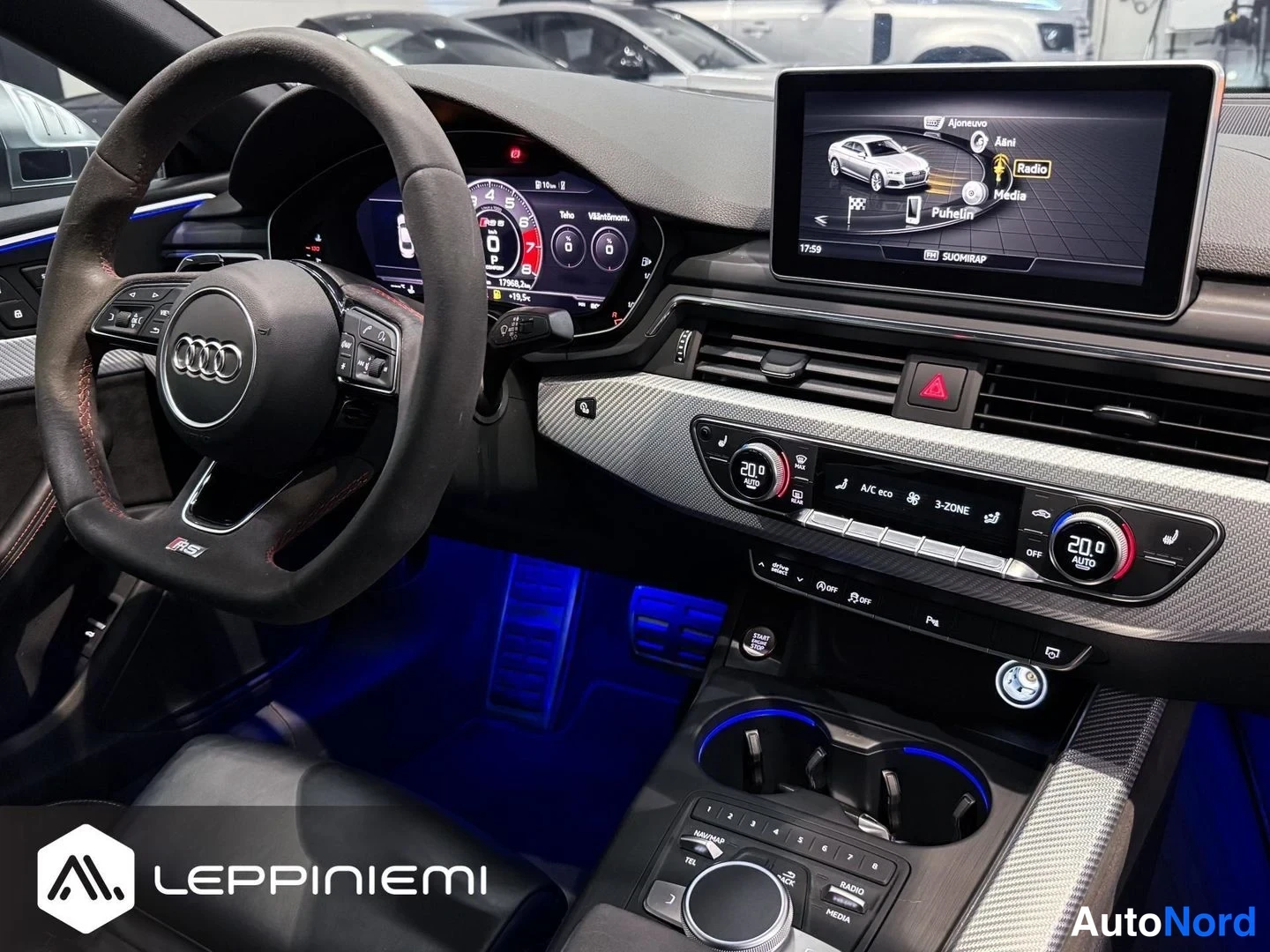 Muu: Ohjaamo lähikuvassa: Audi-ohjauspyörä, digitaalinen mittaristo, keskinäyttö radiolla ja puhelinvalikolla, kaksialueinen ilmastointi 20 astetta.