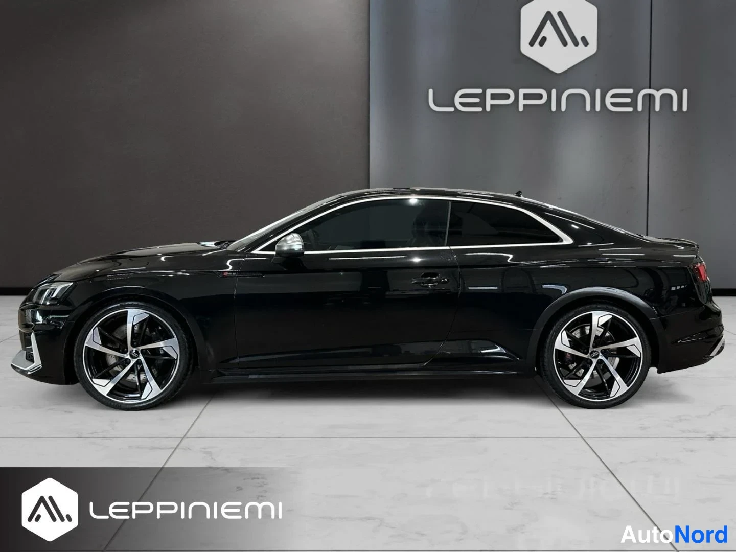Etuviisto, nokka vasemmalle: Mustan coupé-auton sivuprofiili showroomissa, keula lievästi vasemmalle kääntyneenä, harmaa-helmet­kat­kaisu­vanteet.