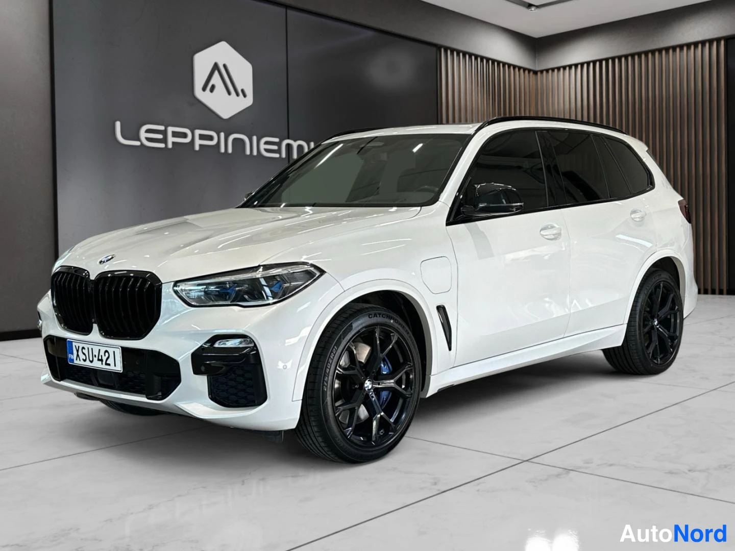 Etuviisto, nokka vasemmalle: Valkoinen BMW X5 kolmeneljäsosan etukuvassa, etusivu ja vasen kylki näkyvissä, mustat 21 tuuman kevytmetallivanteet ja siniset jarrusatulat, rekisterit
