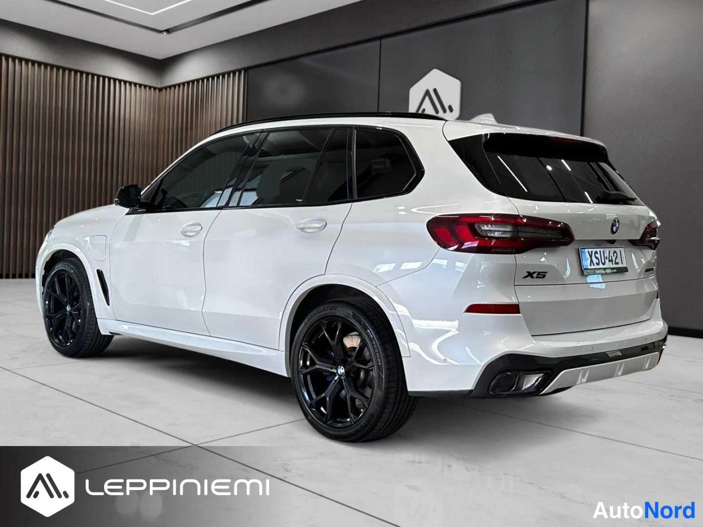 Takaviisto: Valkoisen BMW X5:n takakolmasosa oikealta, mustat vanteet ja siniset jarrusatulat näkyvissä, rekisteritunnus, korissa latausluukku.