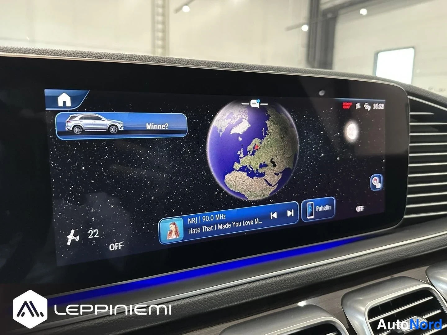 Yksityiskohta: Mercedes-Benzin kojelaudan suuri näyttö, jossa näkyy navigaation maapallonäkymä ja radiokanava NRJ 90,0 MHz.