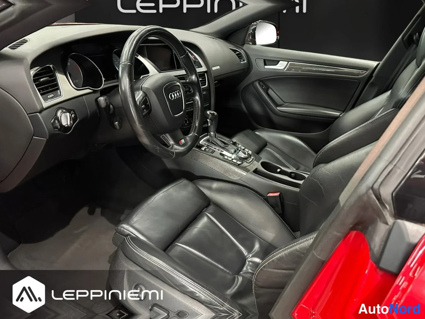 Muu: Auton sisätilan kuva kuljettajan puolelta: musta nahkapinnoitteinen ohjaamo, Audi-ohjauspyörä, automatic-vaihdekeksi ja kaksi etuistuinta.