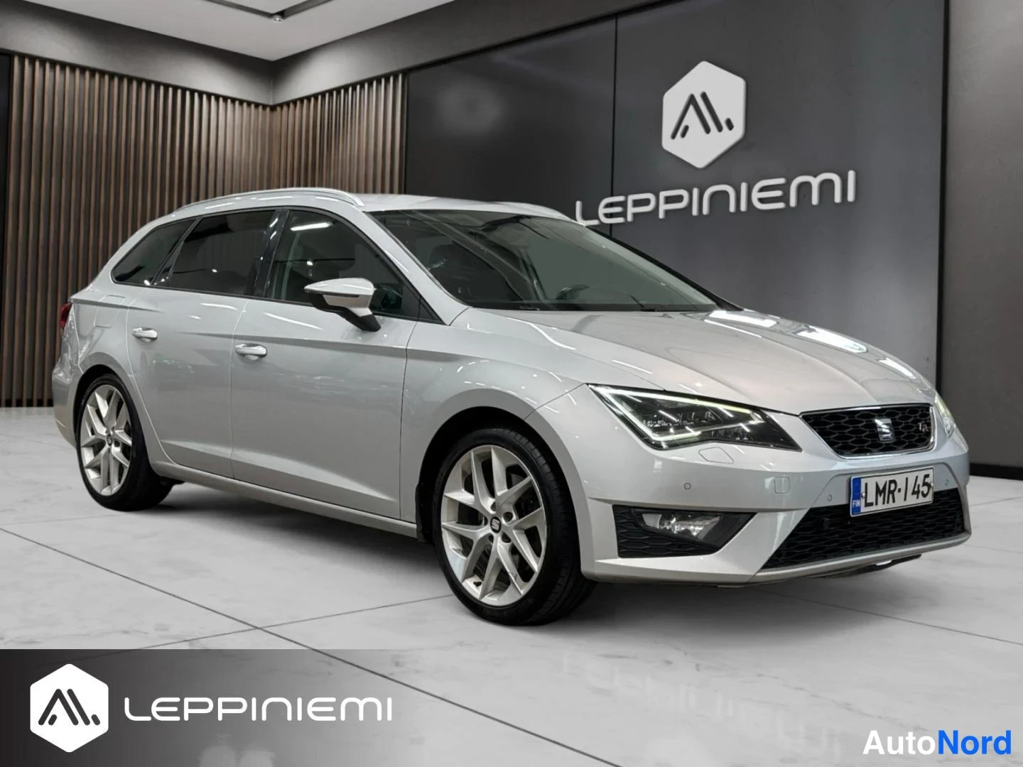 Etuviisto, nokka oikealle: Hopeanvärinen Seat Leon ST -farmariauto esillä myyntihallissa, kolmen neljäsosan etunäkymä oikealta; näkyvissä kylki, etusäleikkö, LED-ajovalot ja rekist