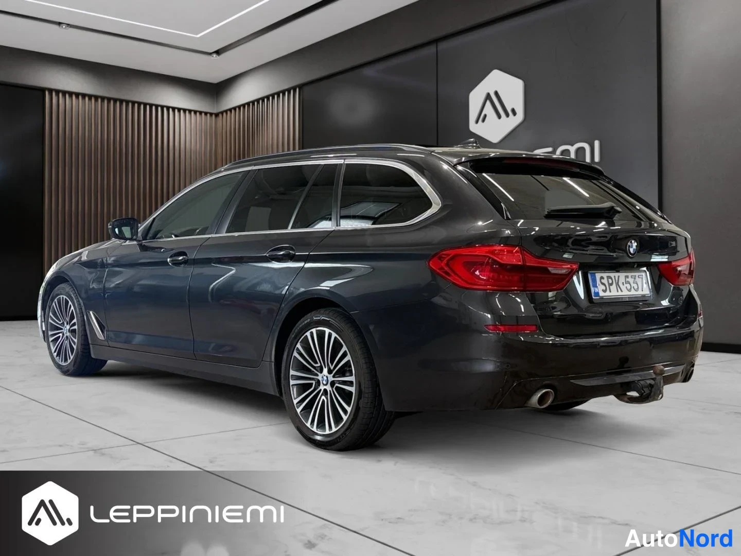 Takaviisto: Tummanharmaa BMW 5-sarjan farmari kuvattuna takaa vasemmasta kulmasta autosalon lattialla; rekisterikilpi, savukehyskatto ja moni literatura-kevytmetallivanteet