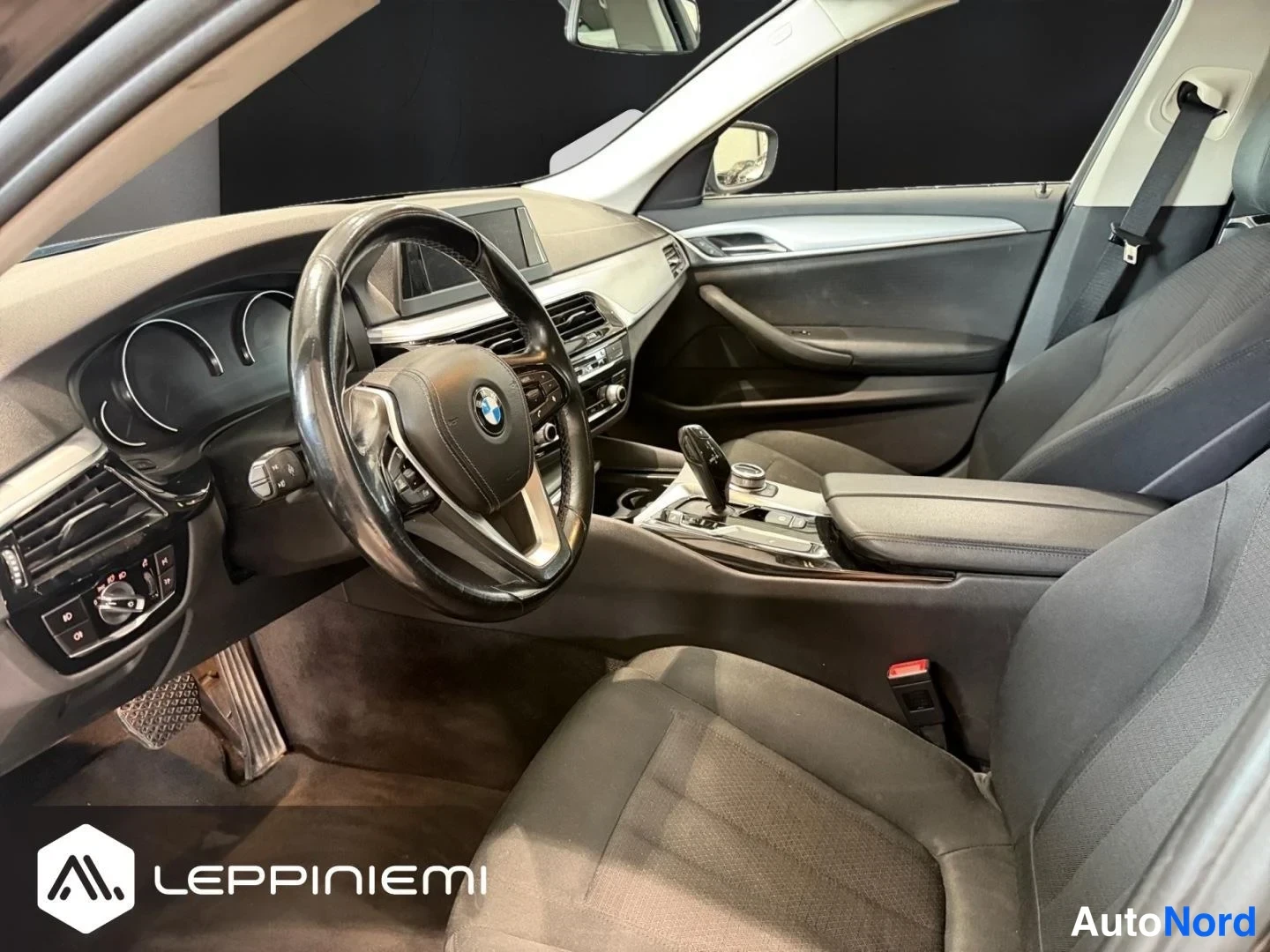 Muu: Sisätilakuva BMW:n etupenkiltä: ohjauspyörä, kojetaulu ja vaihtajanäyttö, tummat kangasistuimet.
