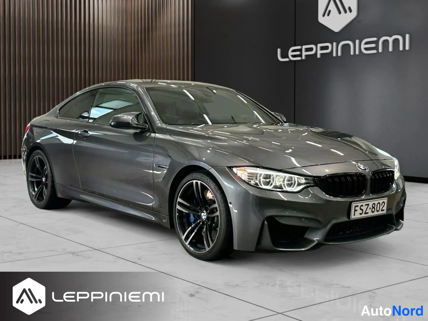 Etuviisto, nokka oikealle: BMW M4 -kupé tummanharmaana kolmannesvärikuvana edessä oikealle kääntyneenä, näkyvät keulaproosi ja kylki, harmaa lattiapinta ja Leppiniemi-tausta.