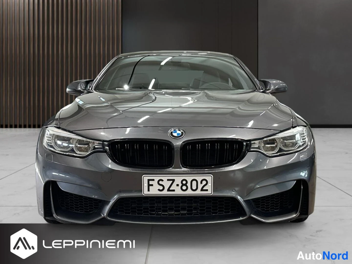 Edestä: BMW M4 -kupé edessäpäin, tummanharmaa konepelti, musta munuaismaski, LED-ajovalot, rekisteritunnus ja harmaat tuolit näkyvät tuulilasin läpi.