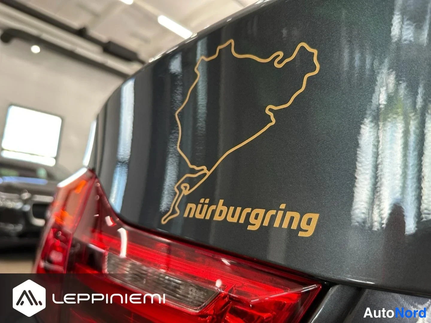 Takaa: Takaosan lähikuva: harmaassa kannessa kultainen Nürburgring-ratakarttataatto ja teksti nürburgring, osa punaista takavaloa näkyy.