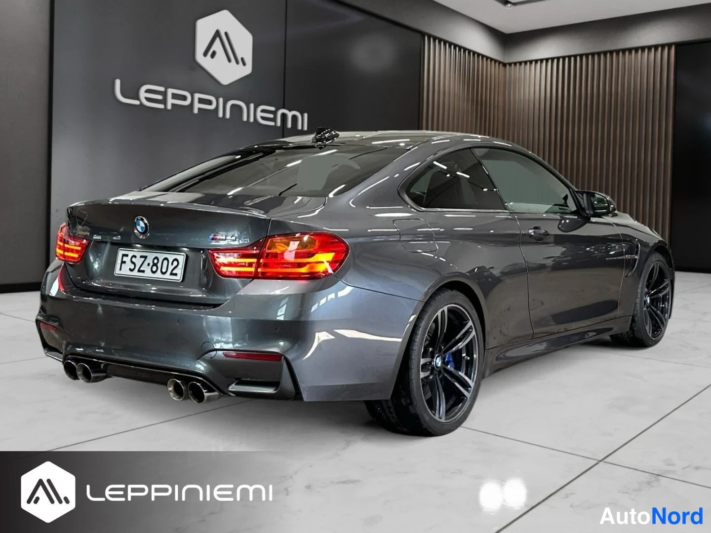 Takaviisto: Auton takakolmannellinen näkymä sisätiloissa: tummanharmaa BMW-coupé, M4-merkintä, rekisteritunnus, neljä pakoputkea ja mustat kevytmetallivanteet.