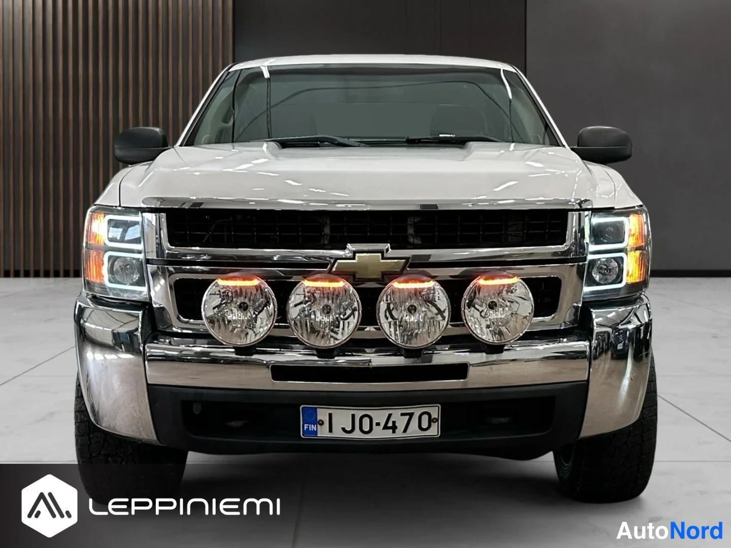 Edestä: Valkoisen Chevrolet Silveradon etupää suoraan edestä, neljä pyöreää lisävaloa ja rekisteritunnus FI.