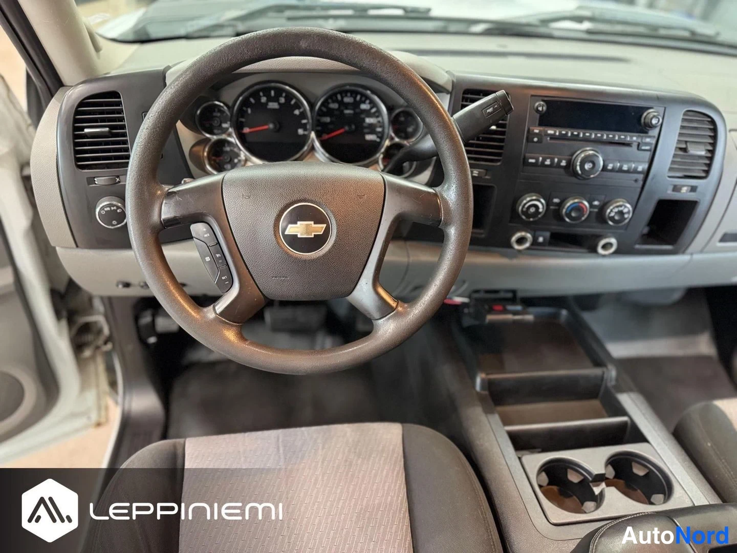 Muu: Ohjaamo näytettynä etusisältä: Chevrolet-merkillinen ohjauspyörä, instrumentit ja radio-konsoli, kangasistuin etualalla.