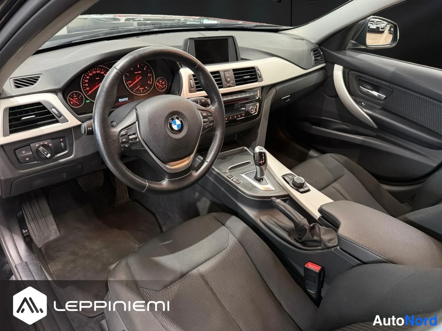 Takaviisto: Ohjaamo kuljettajan puolelta takaa päin: BMW-ohjauspyörä, analogiset mittarit, näyttö keskiöössä ja mustat penkit.
