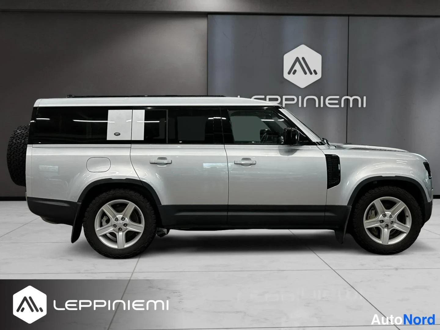 Sivulta: Land Rover Defenderin sivukuva vasemmalta puolelta, hopeinen koripinta, takaan sijoitettu varapyörä ja hopeavanteet.