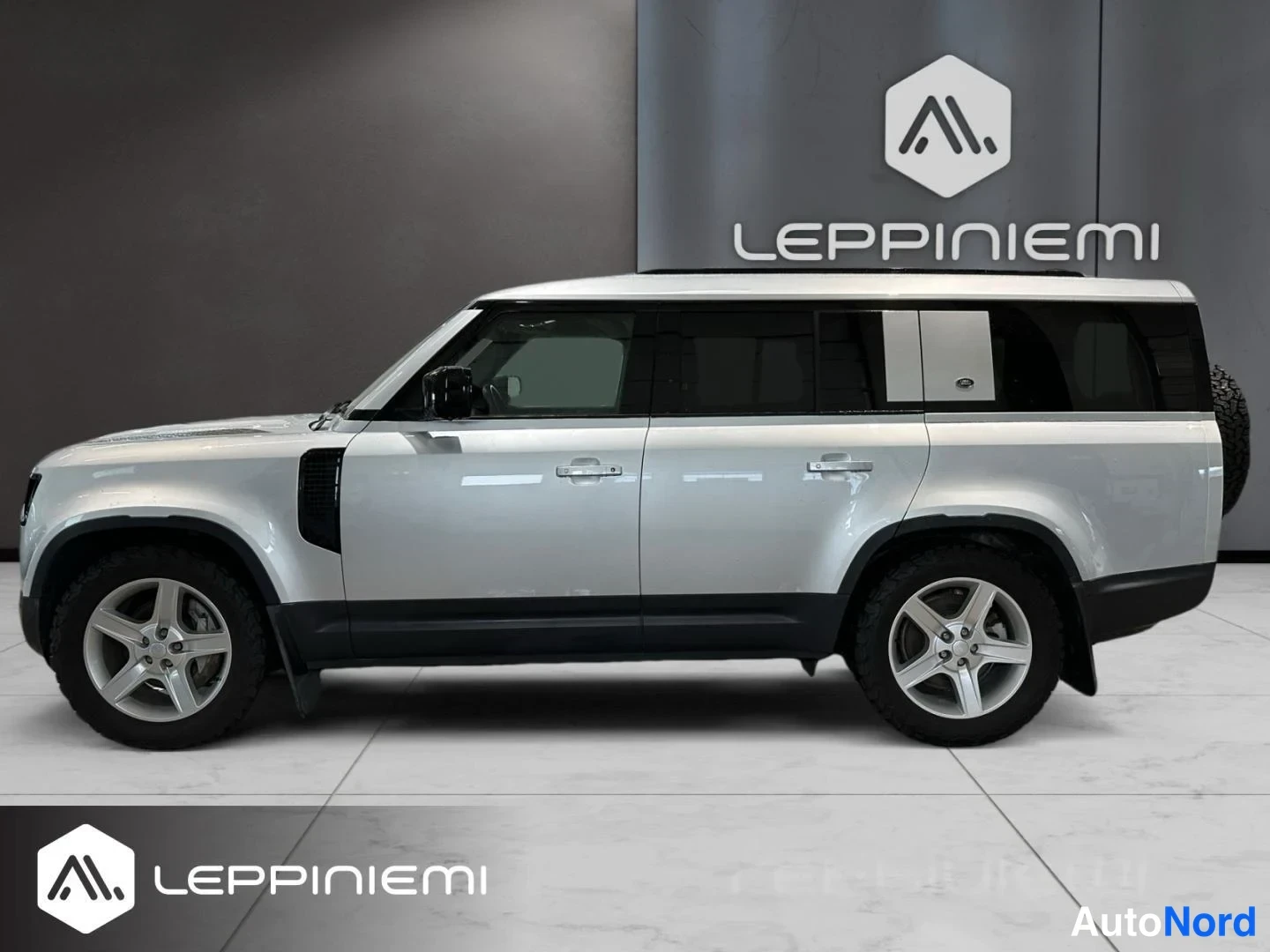 Sivulta: Land Rover Defenderin sivuprofiili: pitkä koriversio, hopeanvärinen kori, tummat ikkunat ja alumiinivanteet autonäyttelyssä.