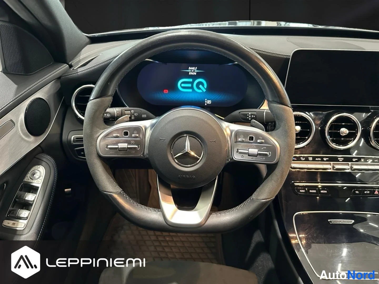 Edestä: Mercedes-Benzin ohjaamo: ratissa merkki, mittaristossa EQ-aloitusruutu ja mediapaneelin ilmastointiaukot.