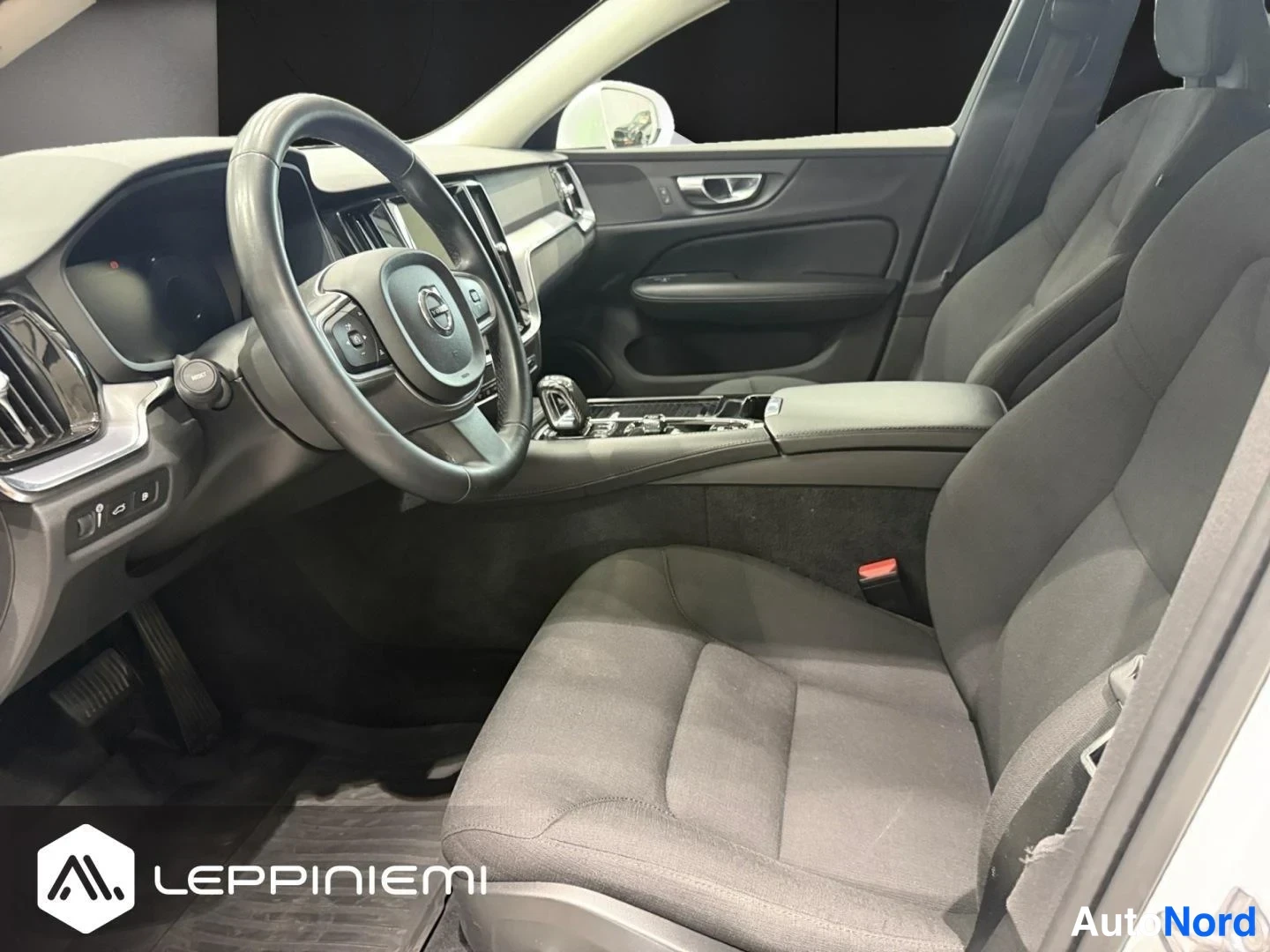 Muu: Volvo V60:n etuistuin ja kojelauta sivulta: musta ohjauspyörä Volvon logolla, keskikonsolin vaihtajan ja näytön ympärillä kiiltävää panelia, tummat kankipenkit.