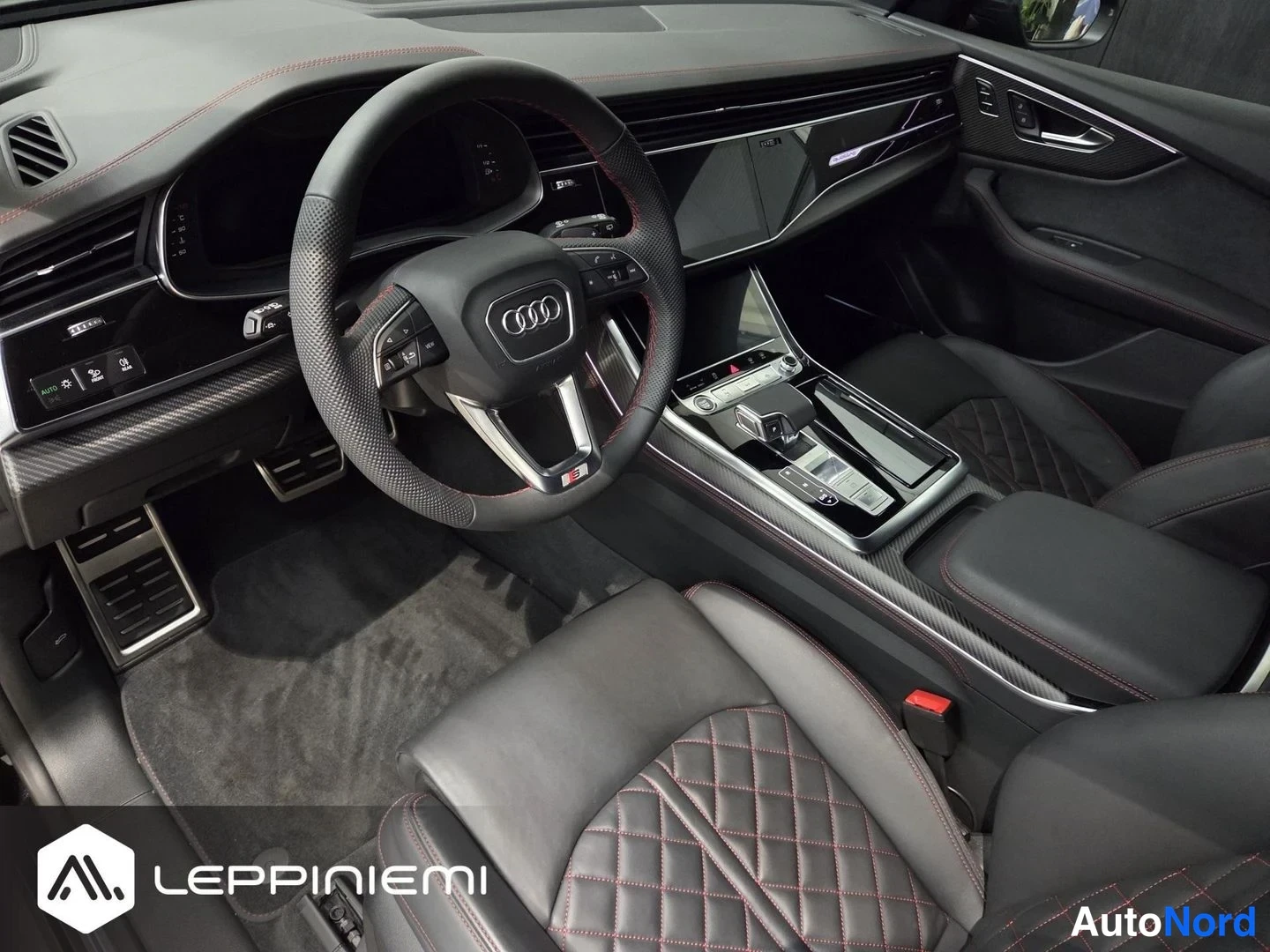 Muu: Audi Q8:n matkustamon sisäkuva: Audi-ohjauspyörä, kosketusnäytöt, käytävä ja rombikuvioiset mustat penkit punaisilla ompeleilla.