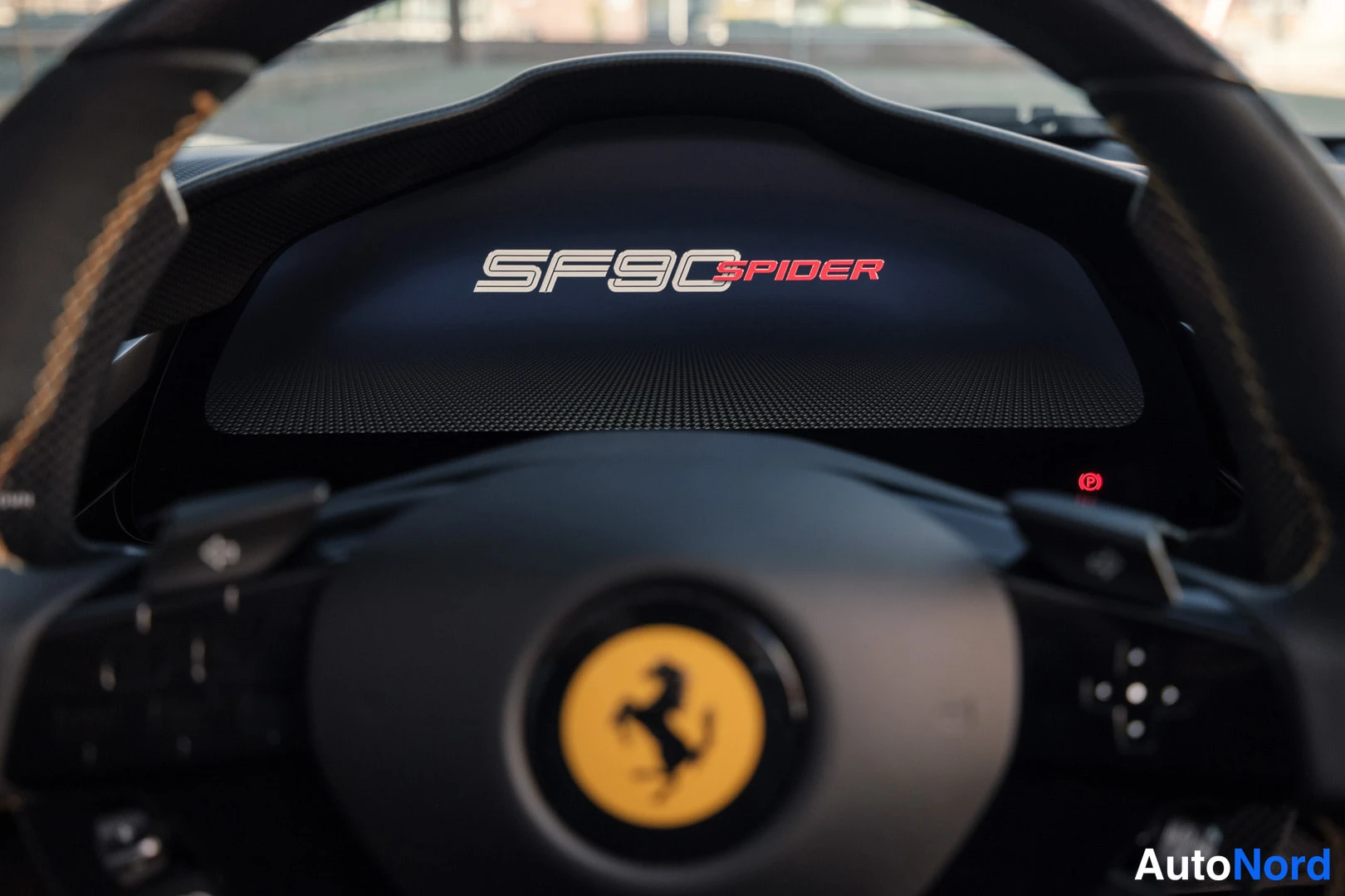 Yksityiskohta: Lähikuva mittarinäytöstä, jossa lukee SF90 SPIDER; etualalla Ferrarin vaakalla varustettu ohjauspyörä.