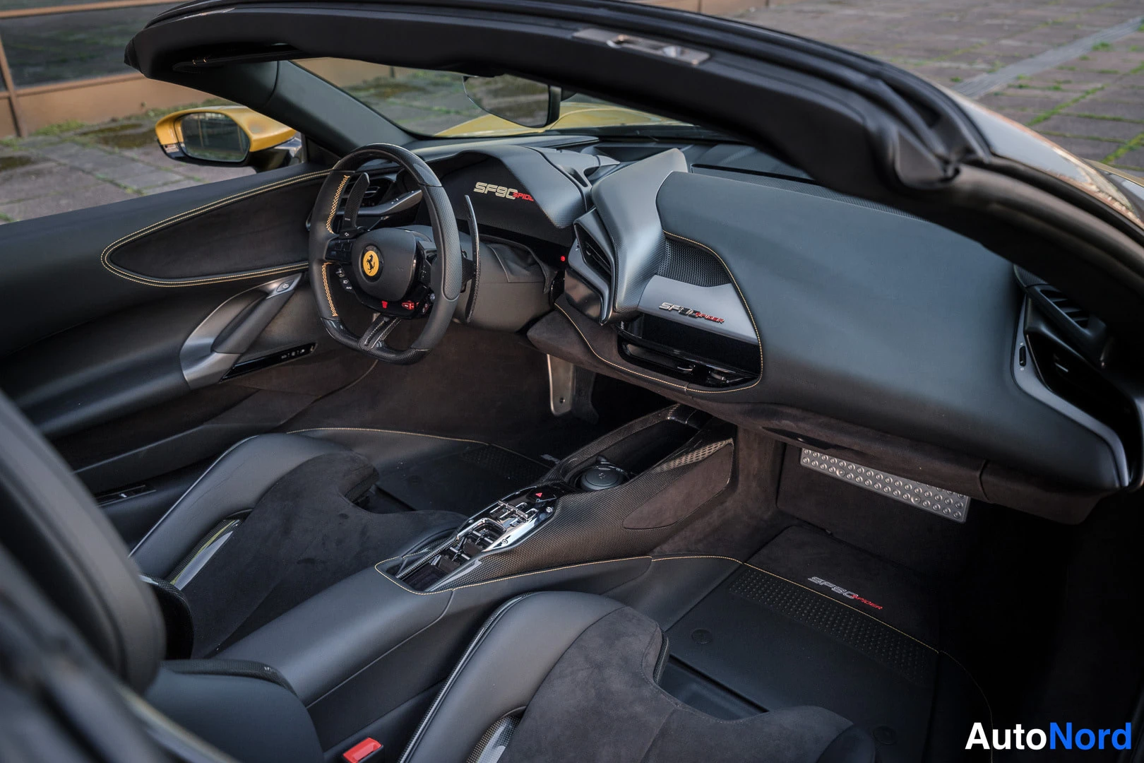 Yksityiskohta: Avo-auton matkustamo ohjaamon puolelta: Ferrari-ohjautumaton ohjauspyörä, mittaristössä teksti 'SF90', keskikonsoliin satunnaisia lukuja, mustat istuimet.
