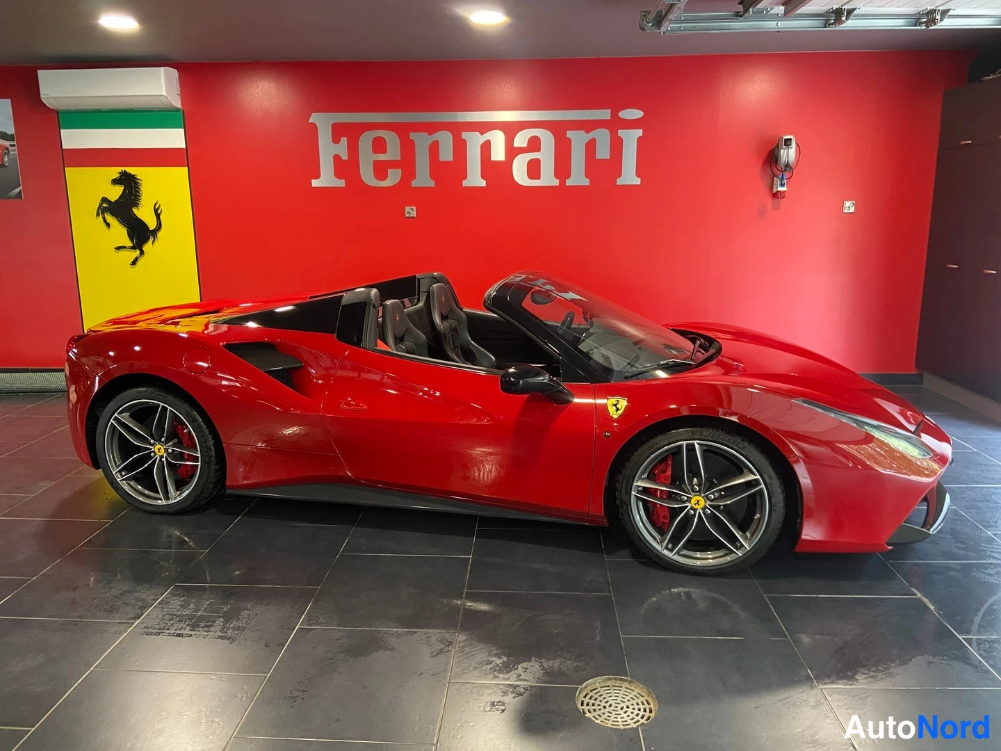 Sivulta: Punainen avokattoinen urheiluauto sivulta kuvattuna autotallissa, jossa punainen seinä ja Ferrari-kyltti. Avokatto ja kaksi istuinta näkyvissä.