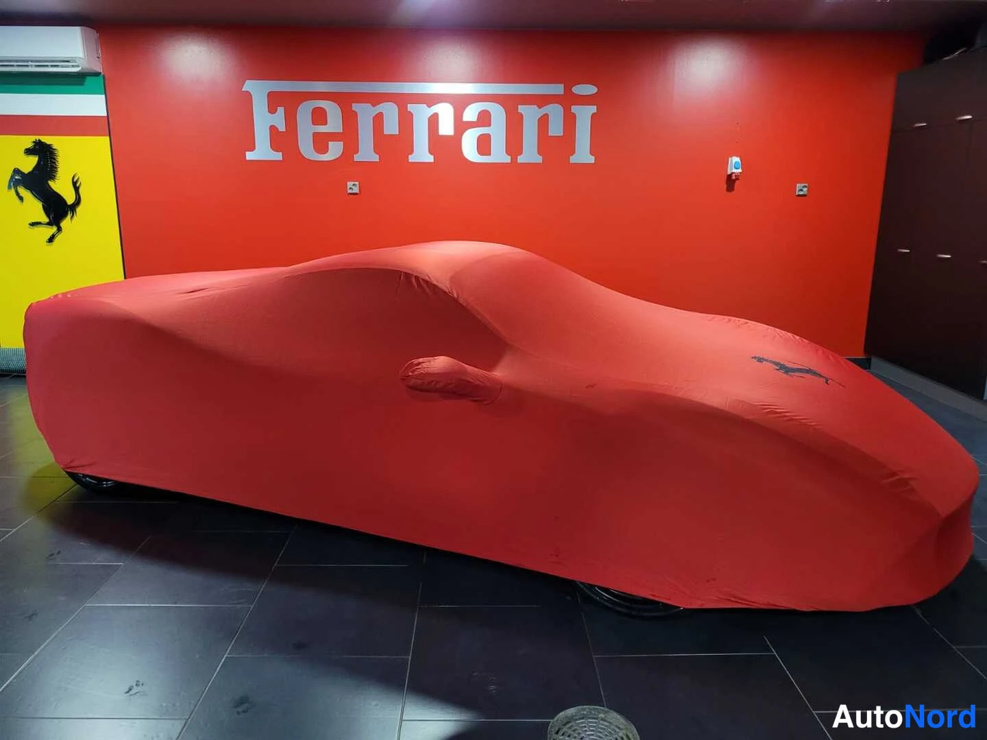 Muu: Punainen Ferrari-talliteltu peittää auton kokonaan lattiassa; taustalla oranssi seinä ja metallinen Ferrari-logo.