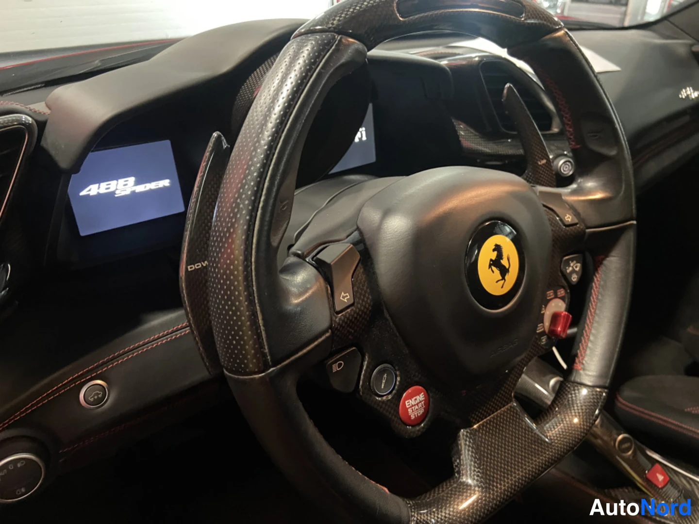 Muu: Lähikuva Ferrari-ohjauspyörästä hiilikuitureunoilla, keskellä keltainen Ferrari-logo ja punainen ENGINE START STOP -painike; ryhtymittarissa lukee '488 SPIDER'.