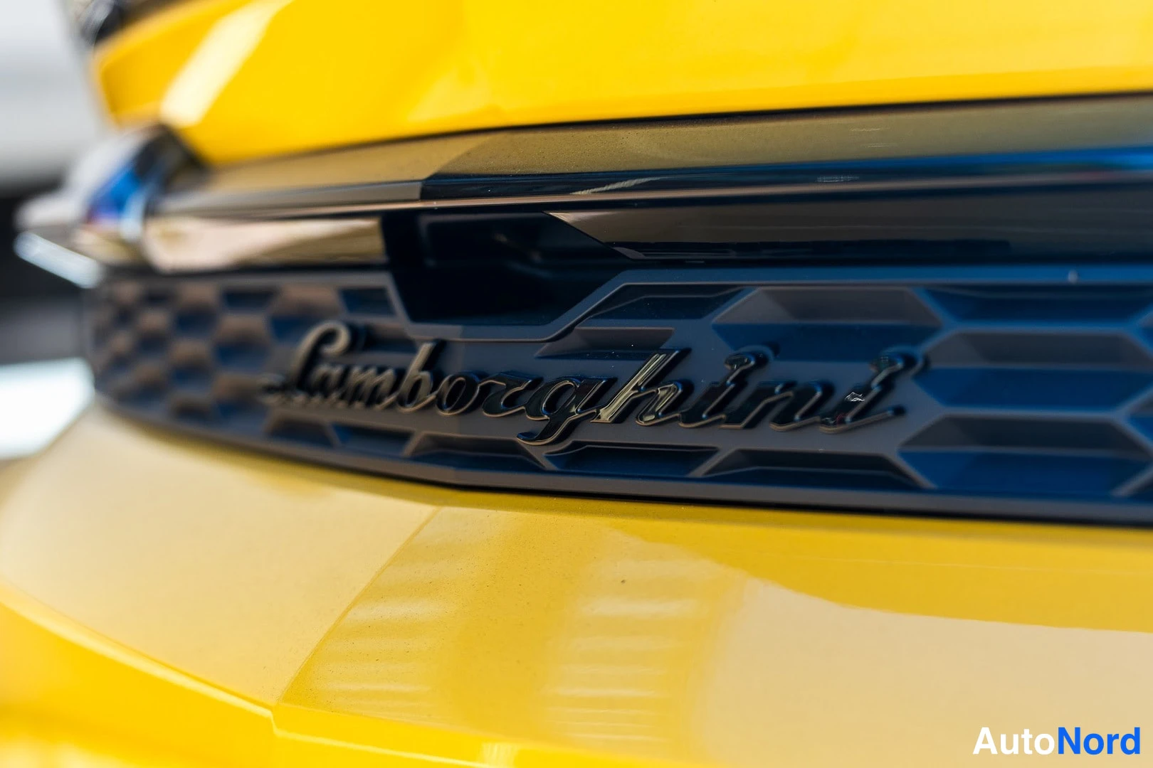 Yksityiskohta: Lähikuva mustasta honeycomb-ritilästä, jossa lukee Lamborghini-kirjoitus, keltaisella koripaneelilla.