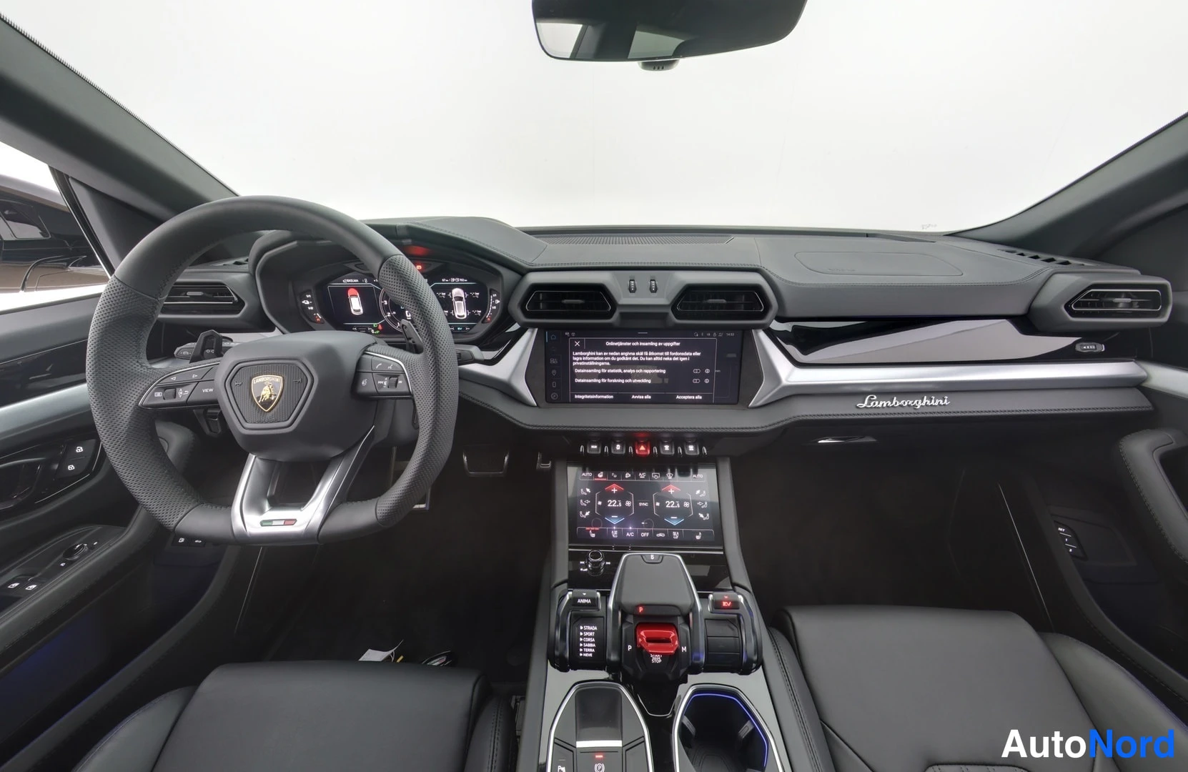 Etuviisto: Ohjaamo suoraan edestä: Lamborghini-ohjauspyörä, digitaalinen mittaristo, keskikonsolin näytöt ja mustat penkit.