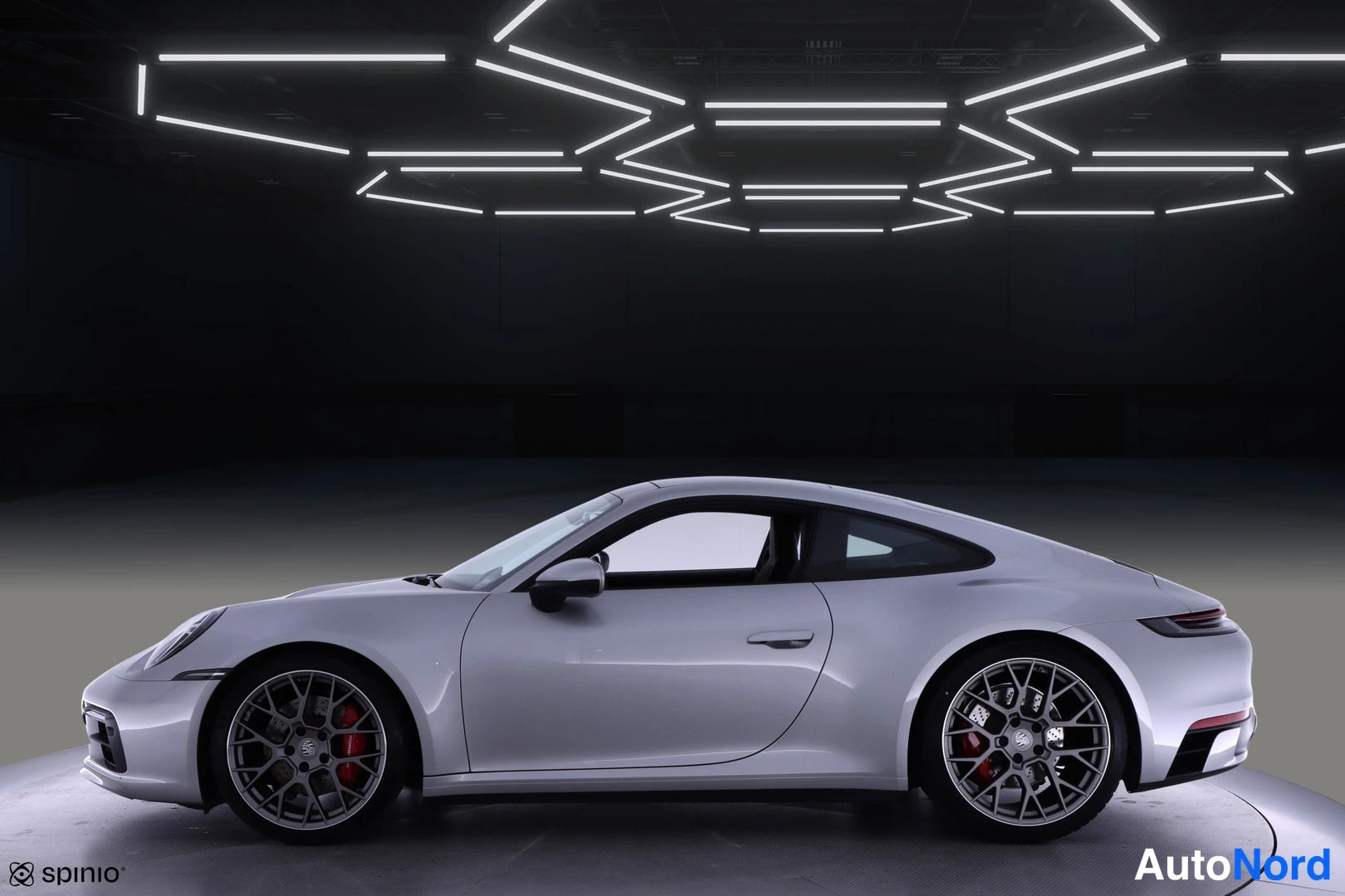 Sivulta: Porsche 911 -auton sivuprofiili; hopeinen coupé, punaiset jarrusatulat ja monisakaraiset kevytmetallivanteet näkyvät tummassa studiossa.