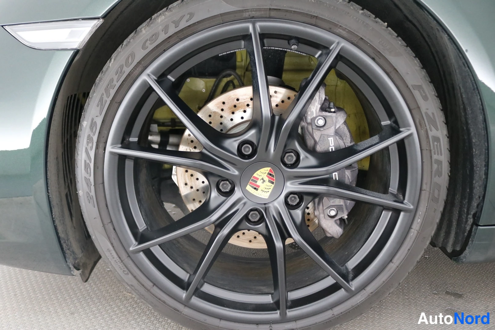 Yksityiskohta: Porsche-väline lähikuva: musta 20 tuuman alumiinivanne, Porsche-merkki navassa ja punaisen/mustan ritiläinen jarrulevy. Renkaan sivuseinässä lukee 245/35 ZR20 (