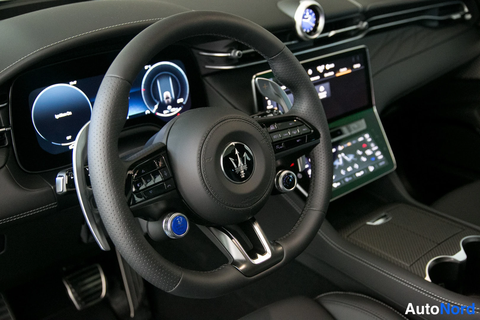 Muu: Lähikuva Maserati-ohjauspyörästä ja kojelaudasta: sininen käynnistyspainike, puhallinvaihdekipät, digitaalinen näyttö ja keskikonsolin säädöt.