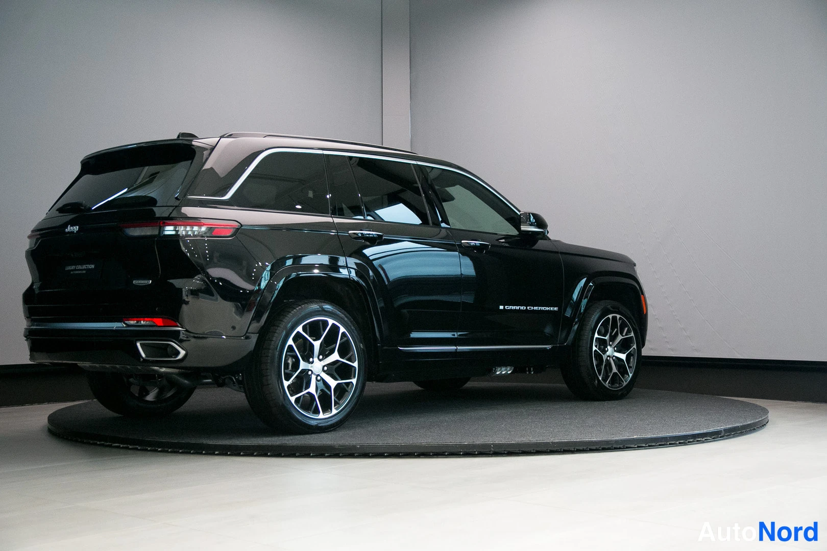 Takaviisto: Musta Jeep Grand Cherokee takaa-vinoalta näkyvä showroomin pyörätelineellä, vasen sivu ja takamootti näkyvissä.