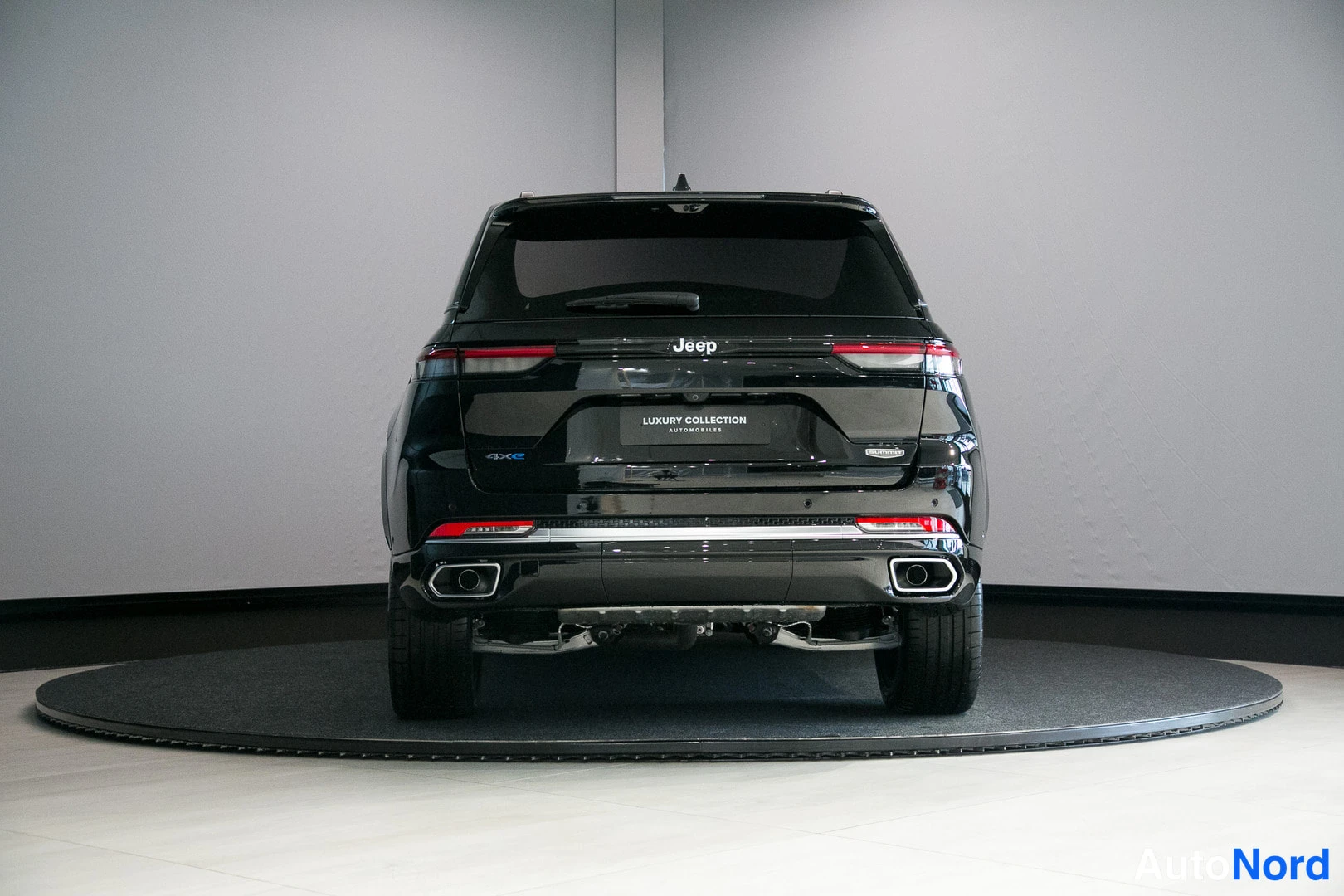 Takaa: Musta Jeepin takanäkymä showroomissa: Jeep-teksti, perävaunut, kaksi pakoputkea ja alustan alla näkyvä tekniikka pyörätelineellä.